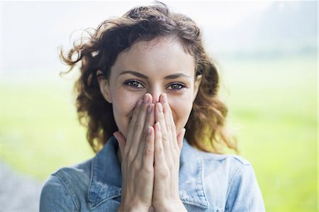 A young woman in a rural landscape, with windblown curly hair. Covering her face with her hands, and laughing. Stock Photo - Premium Royalty-Free, Code: 6118-07235183