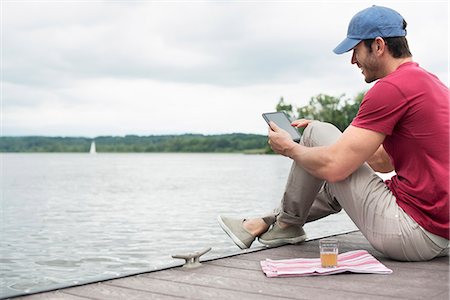 A man seated on a jetty by a lake, using a digital tablet. Stockbilder - Premium RF Lizenzfrei, Bildnummer: 6118-07351269