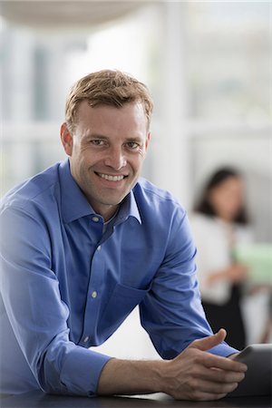 simsearch:6118-07351429,k - Young professionals at work. A man in an open necked shirt using a digital tablet. A group of men and women in the background. Stock Photo - Premium Royalty-Free, Code: 6118-07351360