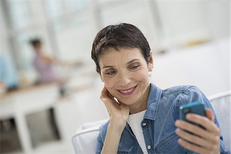 simsearch:6118-07351273,k - Professionals in the office. A light and airy place of work. A mature woman in a blue denim shirt looking at a blue smart phone. Photographie de stock - Premium Libres de Droits, Code: 6118-07351471