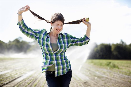 A girl in a green checked shirt with braids standing in a field with sprinklers working in the background. Stock Photo - Premium Royalty-Free, Image code: 6118-07351664