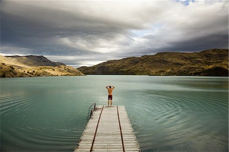 A young man standing at the end of a wooden pier, preparing to dive into calm lake surrounded by mountains in Torres del Paine National Park, Chile. Photographie de stock - Premium Libres de Droits, Code: 6118-07351694