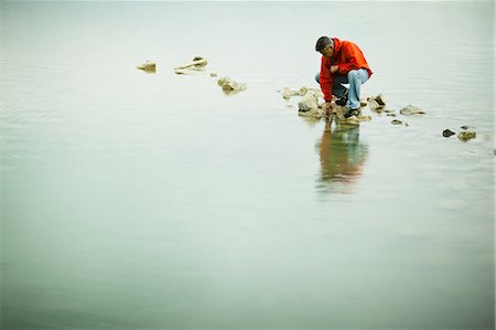 simsearch:6118-07351879,k - A man in a red jacket balanced in a contemplative pose on a stepping stone or rock in shallow water. Stock Photo - Premium Royalty-Free, Code: 6118-07351881