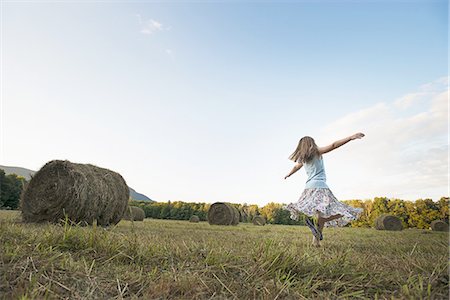 pov - A field full of tall rounded hay bales, and a young girl dancing with her arms outstretched on the stubble field. Stock Photo - Premium Royalty-Free, Code: 6118-07352035
