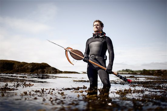 A man in a wetsuit, standing on the shore with a large spear fishing harpoon. Stock Photo - Premium Royalty-Free, Image code: 6118-07352147