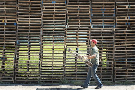 A man carrying a wooden plant frame or pallet in front of a huge stack of objects. Photographie de stock - Premium Libres de Droits, Code: 6118-07352101