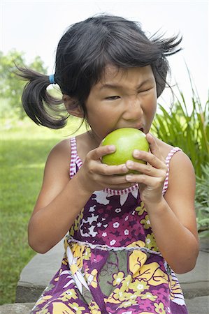 A small child with pigtails chewing a large green apple. Stock Photo - Premium Royalty-Free, Code: 6118-07352337