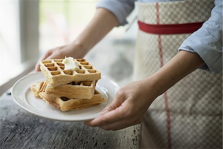 A woman holding a plate of fresh cooked waffles, with cream on top. Stock Photo - Premium Royalty-Free, Code: 6118-07352636