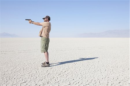 A man aiming a hand gun, holding it with his arm outstretched, standing in a vast, barren desert. Foto de stock - Sin royalties Premium, Código: 6118-07352755