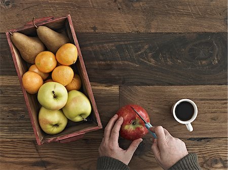 A wooden table top with variety of wood colour and grain. A box of fresh fruits, pears and oranges. A person using a knife to chop an apple. Photographie de stock - Premium Libres de Droits, Code: 6118-07353496