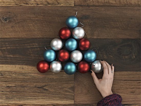 A collection of blue, red and silver ornaments arranged in a triangular shape on a wooden board. A Christmas tree shape. A person's hand placing the final ball. Stock Photo - Premium Royalty-Free, Image code: 6118-07353503