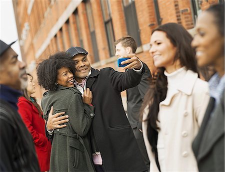 row of people - City life. A group of people on the go. A man holding out a camera phone and taking pictures of the group. Kissing a young woman. Men and women. Stock Photo - Premium Royalty-Free, Code: 6118-07353694