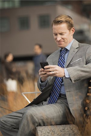 simsearch:6118-07353621,k - A man sitting on a bench outside a large building,  looking at a cell phone screen or mobile phone. Fotografie stock - Premium Royalty-Free, Codice: 6118-07353622