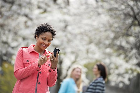 People outdoors in the city in spring time. White blossom on the trees. A young woman checking her cell phone, and laughing. Stock Photo - Premium Royalty-Free, Code: 6118-07354324
