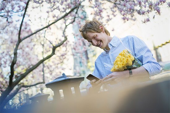 City life. A young man in the park in spring, using a mobile phone.  Holding a bunch of yellow roses. Stock Photo - Premium Royalty-Free, Image code: 6118-07354499