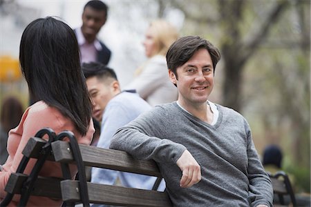 sitting back bench - City life in spring. Young people outdoors in a city park. Sitting on a park bench. A man smiling at the camera. Four people in the background. Stock Photo - Premium Royalty-Free, Code: 6118-07354563