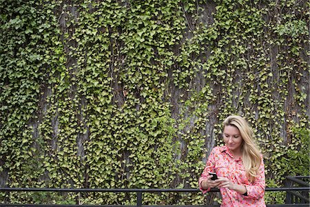 City life in spring. City park with a wall covered in climbing plants and ivy.  A young blonde haired woman checking her smart phone. Stock Photo - Premium Royalty-Free, Code: 6118-07354587
