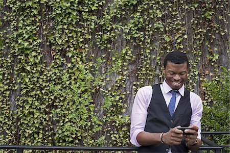 people - City life in spring. City park with a wall covered in climbing plants and ivy.  A young man in a waistcoat, shirt and tie checking his phone. Stock Photo - Premium Royalty-Free, Code: 6118-07354589