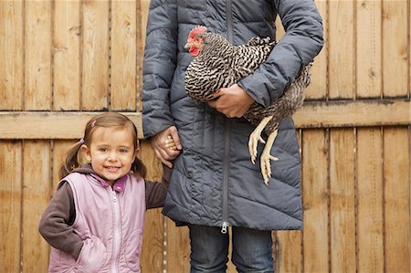 feathers woman - A woman in a grey coat holding a black and white chicken with a red coxcomb under one arm. A young girl beside her holding her other hand. Stock Photo - Premium Royalty-Free, Code: 6118-07354590