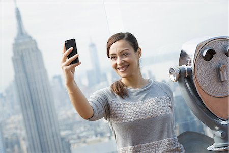 New York City. An observation deck overlooking the Empire State Building. A woman using her smart phone to take a photograph of herself and the view over the city. Stock Photo - Premium Royalty-Free, Code: 6118-07354514