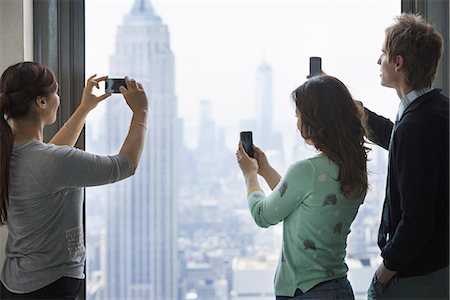 sacar fotos - Urban lifestyle. Three people standing on an observation deck, using their phones to take images of the view over the city. Foto de stock - Sin royalties Premium, Código: 6118-07354522