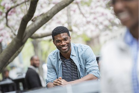 Outdoors in the city in spring. An urban lifestyle. A man sitting at a table smiling. A woman in the foreground. Stock Photo - Premium Royalty-Free, Code: 6118-07354743