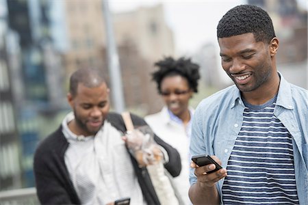 sophisticated - Outdoors in the city in spring. An urban lifestyle. A young man checking his phone and texting. A couple behind him. Stock Photo - Premium Royalty-Free, Code: 6118-07354750