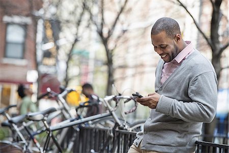 spring season and african american people - Outdoors in the city in spring. An urban lifestyle. A man using his phone. A group of people in the background. Cycle rack. Stock Photo - Premium Royalty-Free, Code: 6118-07354729