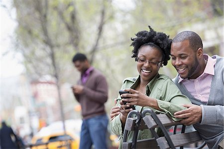 Outdoors in the city in spring. An urban lifestyle. A woman and a man on a bench checking her phone. Two men in the background. Foto de stock - Sin royalties Premium, Código: 6118-07354735