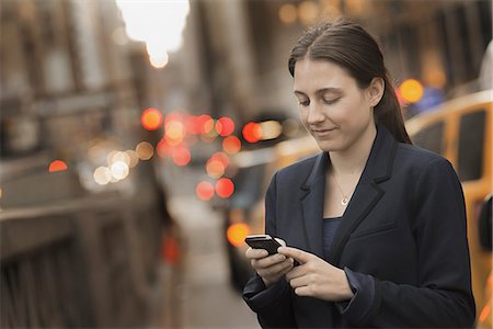 simsearch:6118-07353621,k - A woman in a business jacket checking her cell phone, on a city sidewalk at dusk. Fotografie stock - Premium Royalty-Free, Codice: 6118-07440407