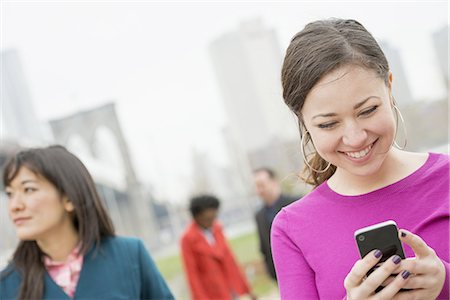 New York city, the Brooklyn Bridge crossing over the East River. Four friends in the park by the river, one woman looking at her phone and smiling. Stock Photo - Premium Royalty-Free, Code: 6118-07440943