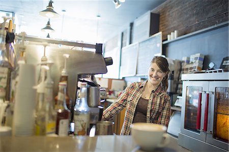 A person, barista, behind the counter at a coffee shop. A large coffee machine for making fresh coffee. A cappuccino in a white cup on the counter. Stock Photo - Premium Royalty-Free, Code: 6118-07439816