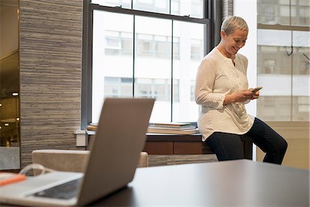 Office life. A woman seated on the edge of her desk using a digital tablet. Stock Photo - Premium Royalty-Free, Code: 6118-07769483