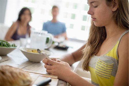 A young girl seated checking her smart phone at a dining table. Two people in the background. Stock Photo - Premium Royalty-Free, Code: 6118-07769557