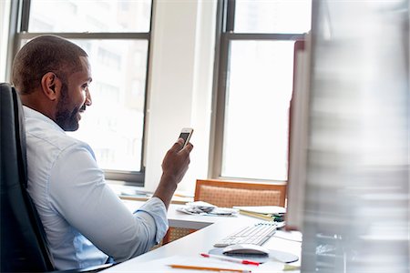 simsearch:6118-07781630,k - A man working in an office seated at a desk, checking his smart phone. Fotografie stock - Premium Royalty-Free, Codice: 6118-07781657