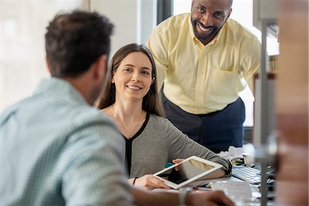 someone sitting at a desk backside - Three people in an office, two men and  a woman with computer monitor and digital tablet. Stock Photo - Premium Royalty-Free, Code: 6118-07781693