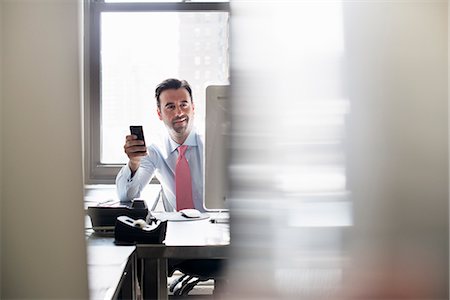 distancia (medición) - A man holding a smart phone, seated at a computer monitor. Foto de stock - Sin royalties Premium, Código: 6118-07781634