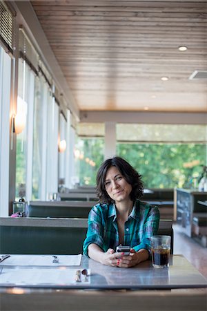 A woman in a checked shirt sitting at a table, laughing and looking at her smart phone. Photographie de stock - Premium Libres de Droits, Code: 6118-07781817