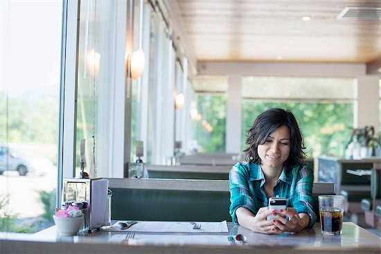 A woman in a checked shirt sitting at a table, laughing and looking at her smart phone. Photographie de stock - Premium Libres de Droits, Le code de l’image : 6118-07781814