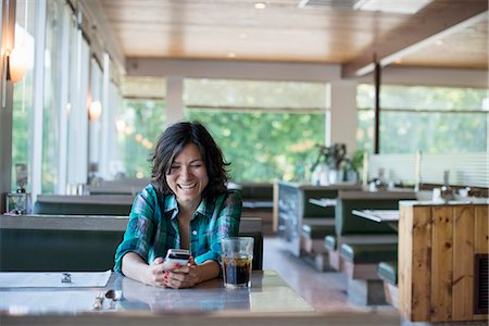 A woman in a checked shirt sitting at a table, laughing and looking at her smart phone. Foto de stock - Sin royalties Premium, Código: 6118-07781816