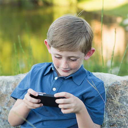 A young boy outdoors sitting leaning against a rock, using a handheld electronic game. Stockbilder - Premium RF Lizenzfrei, Bildnummer: 6118-07732035