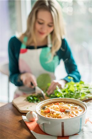 simsearch:6118-07769571,k - A woman wearing an apron, sitting at a table, chopping herbs, a bowl of vegetable stew in the foreground. Fotografie stock - Premium Royalty-Free, Codice: 6118-07966881