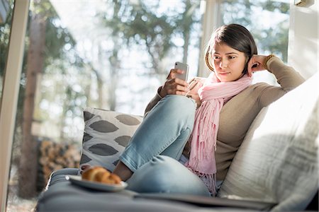 female front - Woman sitting on a sofa looking at her cell phone, a plate with a croissant in front of her. Stock Photo - Premium Royalty-Free, Code: 6118-07966893