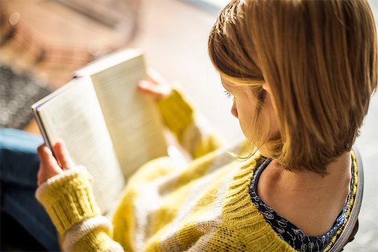 High angle view of a blond girl in a yellow jumper sitting on a chair, reading a book. Stock Photo - Premium Royalty-Free, Image code: 6118-07966896