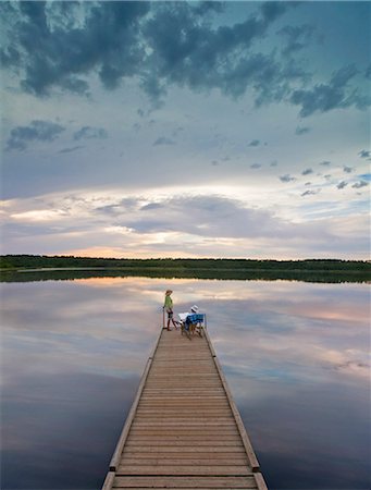 A couple, man and woman sitting at the end of a long wooden dock reaching out into a calm lake, at sunset. Photographie de stock - Premium Libres de Droits, Code: 6118-08001595