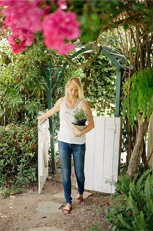 simsearch:6113-06753923,k - Blond woman entering a garden through a white wooden gate, carrying a plant pot. Photographie de stock - Premium Libres de Droits, Code: 6118-08282236