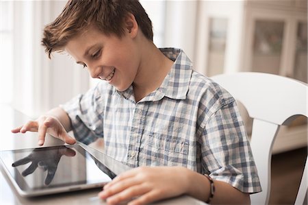 simsearch:6118-08351907,k - A boy seated at a table using a digital tablet, hand on the touch screen. Fotografie stock - Premium Royalty-Free, Codice: 6118-08351894