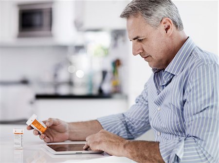 A man seated at a table using a digital tablet, holding a medicine pill bottle, and reading the label. Stock Photo - Premium Royalty-Free, Code: 6118-08393956