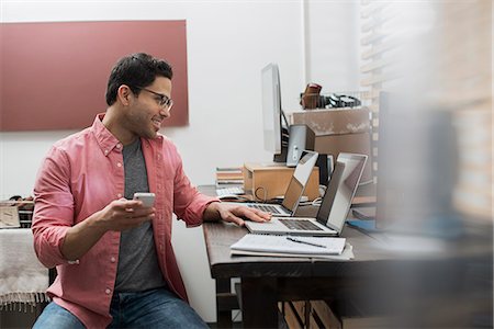 simsearch:6118-07966891,k - A man in a home office with a desk with two laptops, checking information on the screen, holding a smart phone. Photographie de stock - Premium Libres de Droits, Code: 6118-08394068