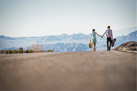 A young couple, man and woman walking hand in hand on a tarmac road in the desert carrying cases. Stock Photo - Premium Royalty-Free, Code: 6118-08394227